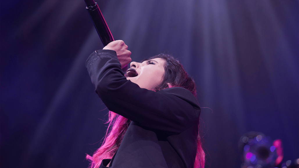 A woman sings K Pop music into a microphone on stage, as a viewer wonders, ‘What is K pop music?’.