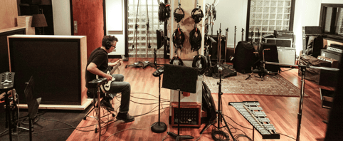 A musician strums on the guitar inside a recording studio filled with instruments.