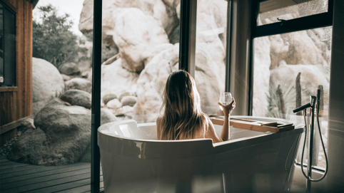 A woman in white bathtub with a view holds up a drinking glass, while listening to bathtub chill songs